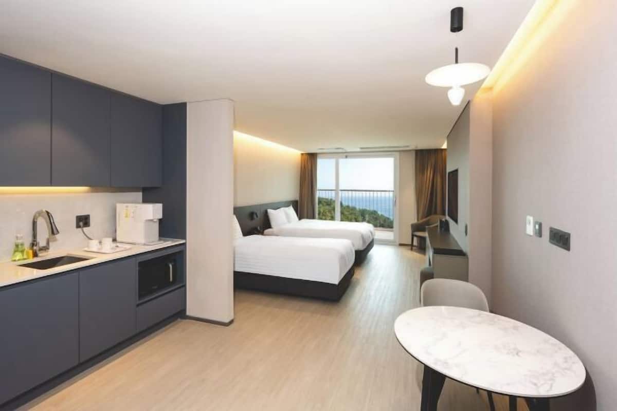 A modern hotel room with a kitchen area, two beds, a dining table, and a balcony with a view.