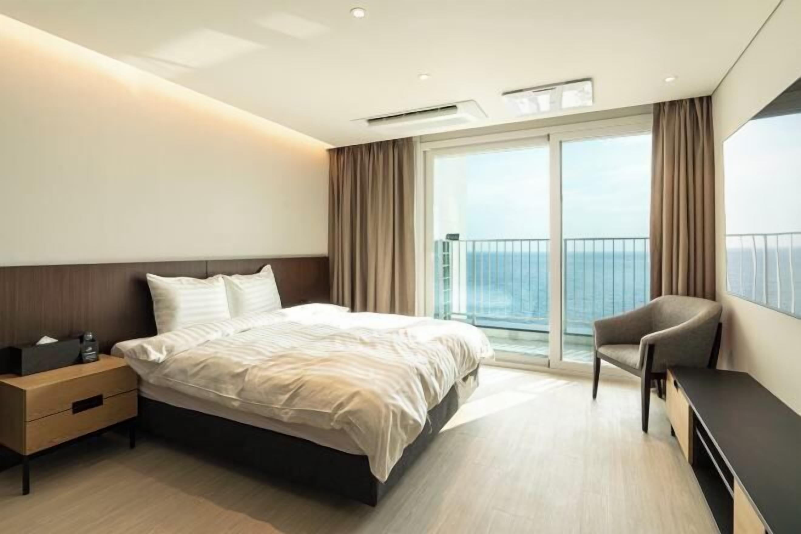 A modern hotel room with a large bed, a bedside table, a chair, and a balcony with a view of the sea.