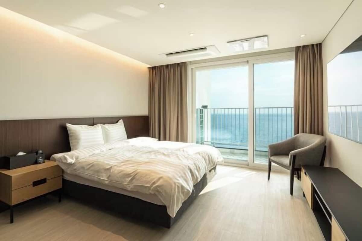 A modern hotel room with a large bed, a bedside table, a chair, and a balcony with a view of the sea.