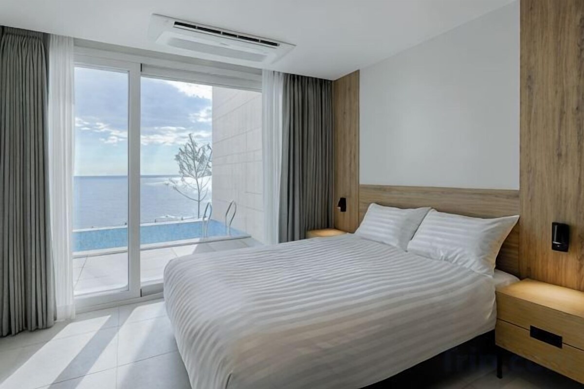 Een moderne slaapkamer met een groot bed, een houten hoofdbord, een nachtkastje en uitzicht op zee door een schuifdeur.