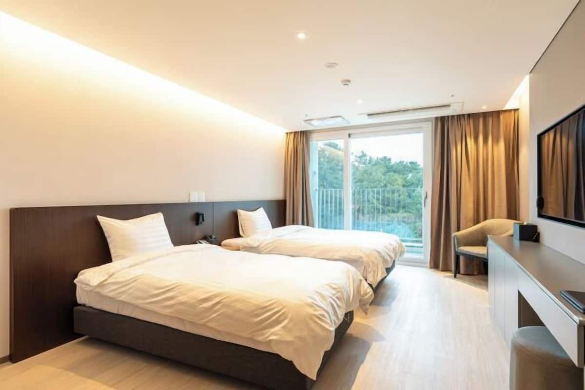 A hotel room with two beds, a desk, a chair, and a large window with curtains.