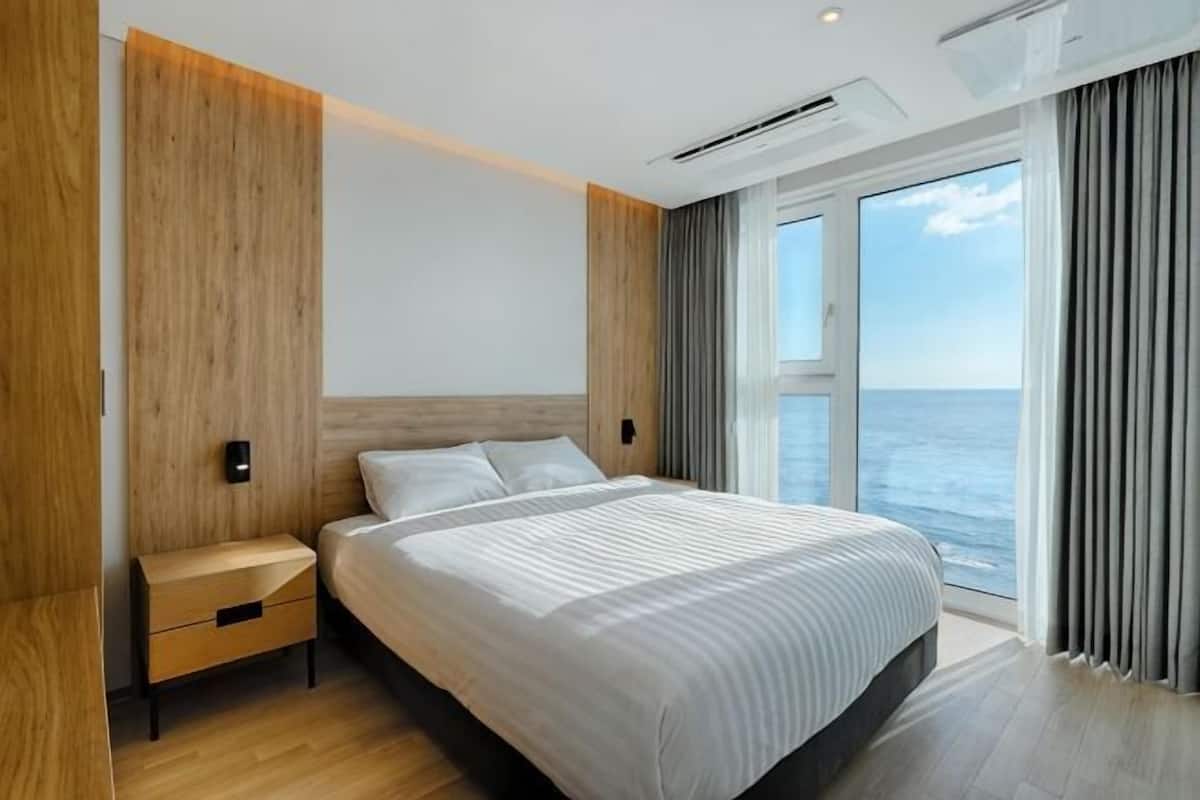 A modern hotel room with a large bed, a nightstand, a wooden accent wall, and a view of the sea.