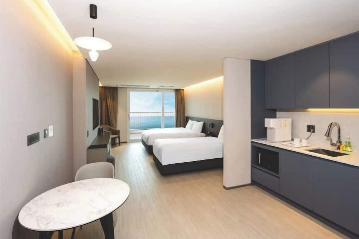 A hotel room with two beds, a dining table, a kitchen area, and a view of the sea.