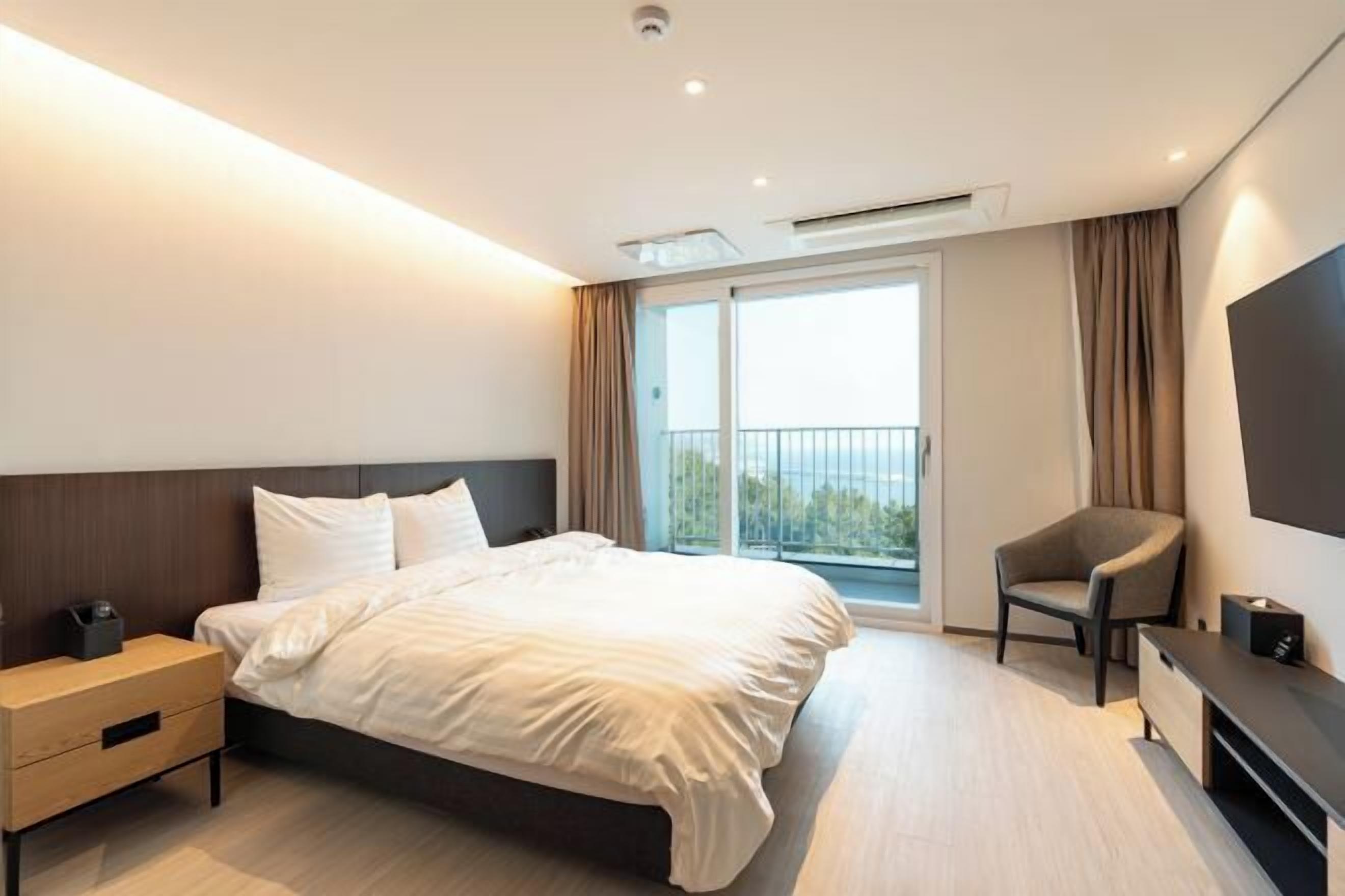 A modern hotel room with a bed, a nightstand, a chair, a TV, and a balcony with curtains.