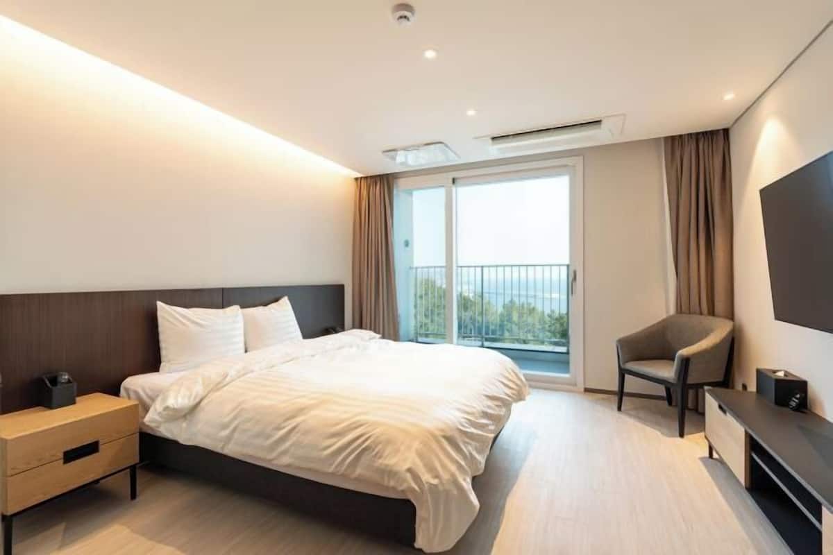 A modern hotel room with a bed, a nightstand, a chair, a TV, and a balcony with curtains.