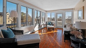 Una habitación luminosa con grandes ventanales, muebles de mimbre y piso de baldosas.