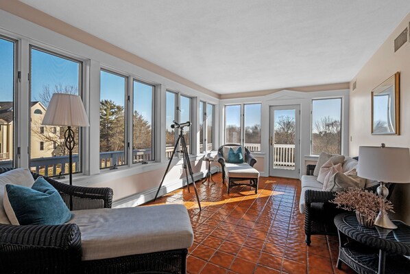 A sunlit room with large windows, wicker furniture, and a tiled floor.