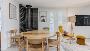 A modern dining area with a round wooden table, six wooden chairs, a yellow armchair, and a black and white brick wall. - Slow M Ocean Cabarita - Pet Friendly (Bogangar)