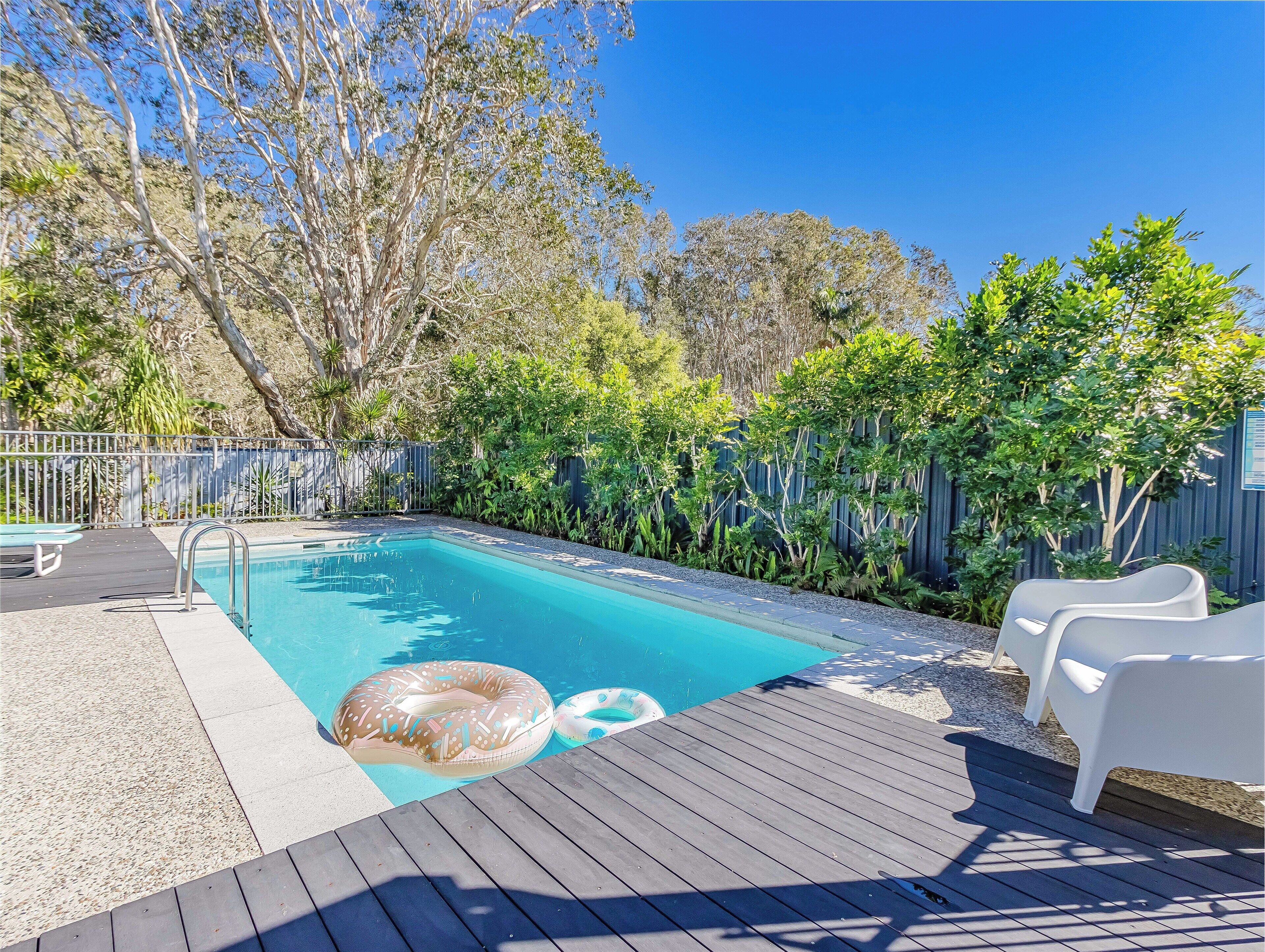 A swimming pool with a wooden deck, surrounded by a fence and various plants.