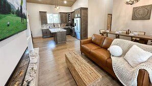 A spacious living area with a brown leather sofa, a woven bench, and a dining table set for four. A kitchen with modern appliances and a large island is visible in the background. - Charming & Luxurious 5-Bedroom Cabin in Heber (Overgaard)