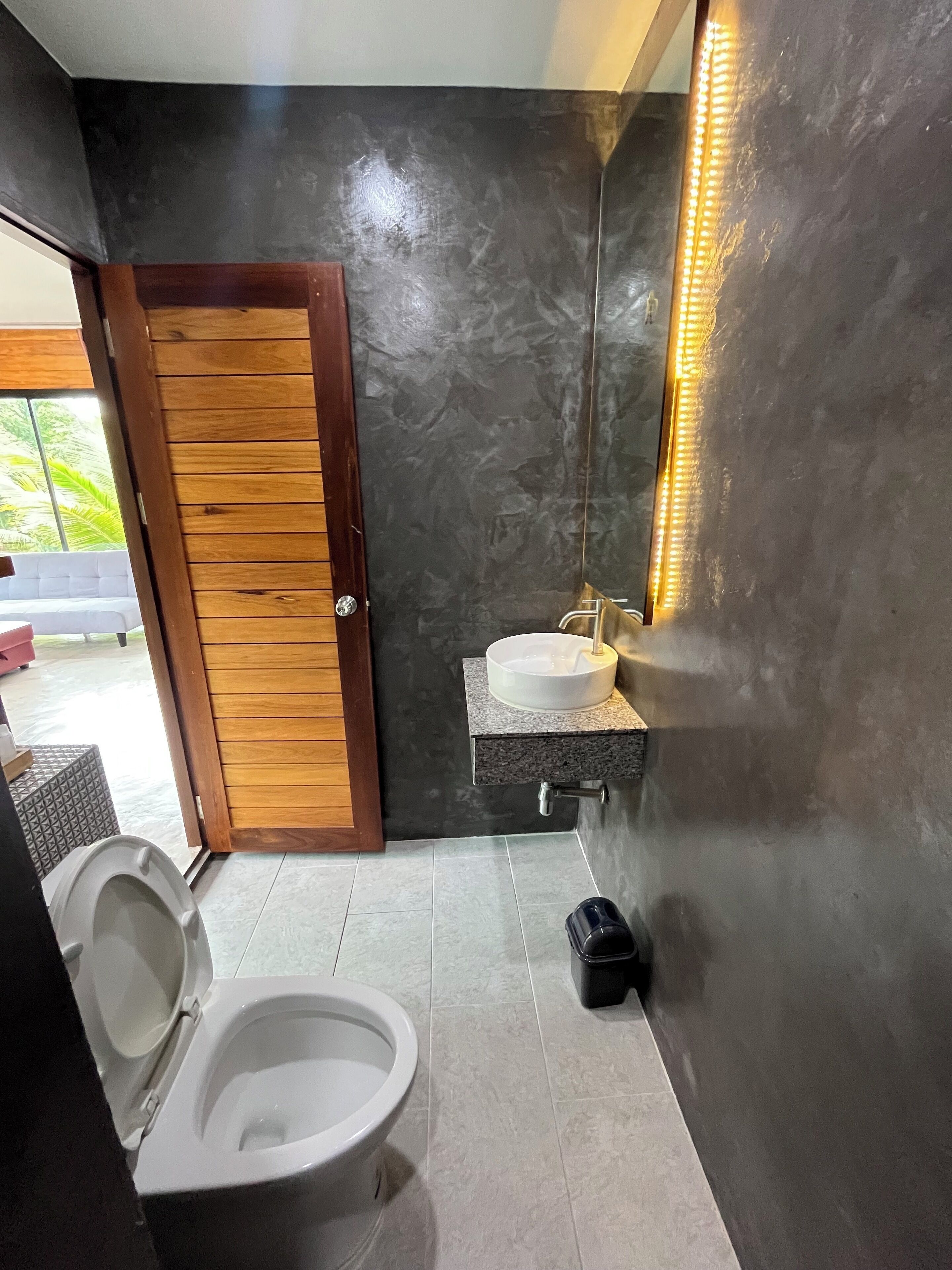 Un bagno moderno con una porta in legno, un water bianco, un lavabo con piano in marmo e uno specchio con illuminazione a LED.