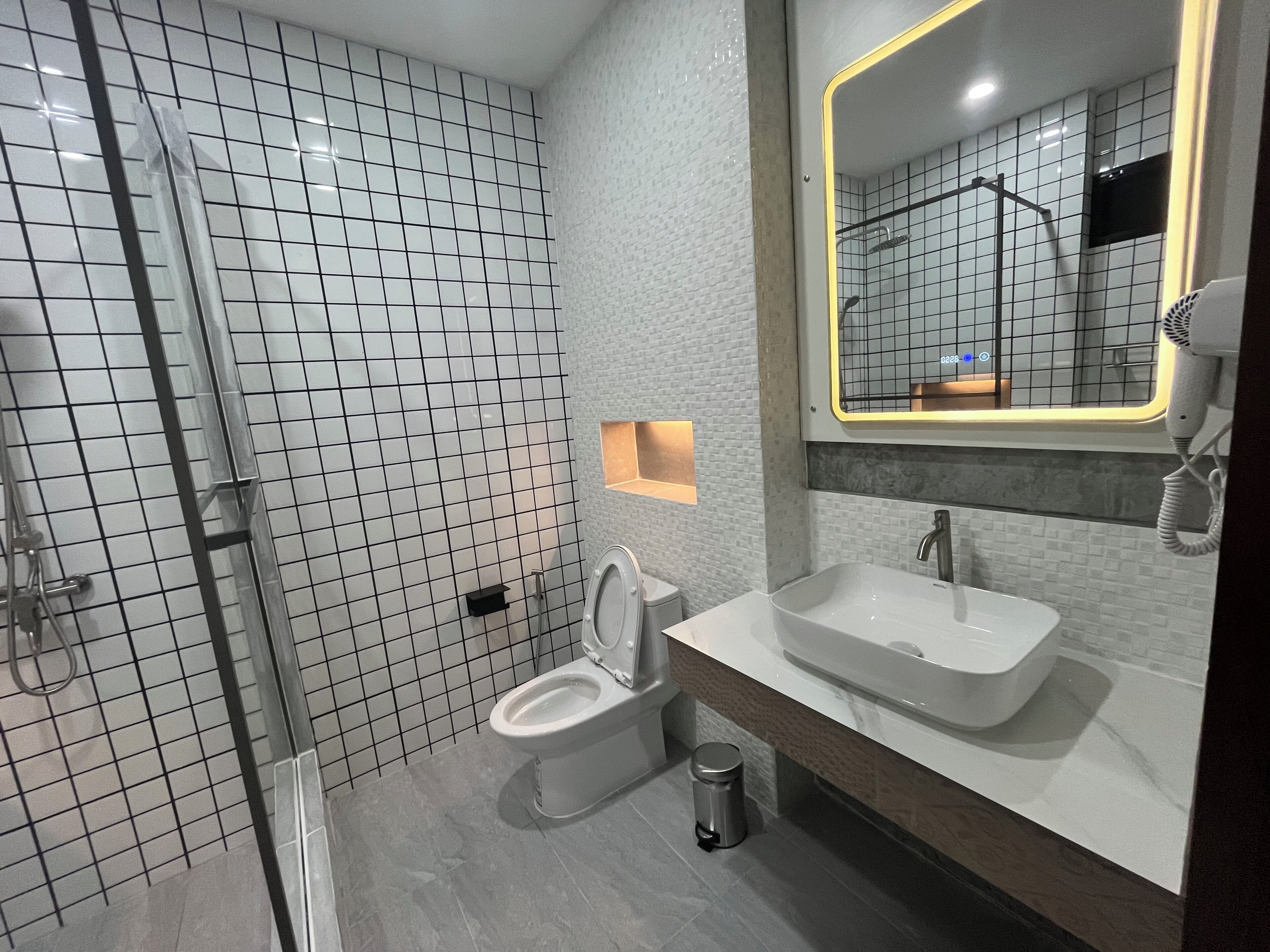 Un baño moderno con ducha, inodoro y lavamanos, que presenta un revestimiento de azulejos y un espejo con marco iluminado.
