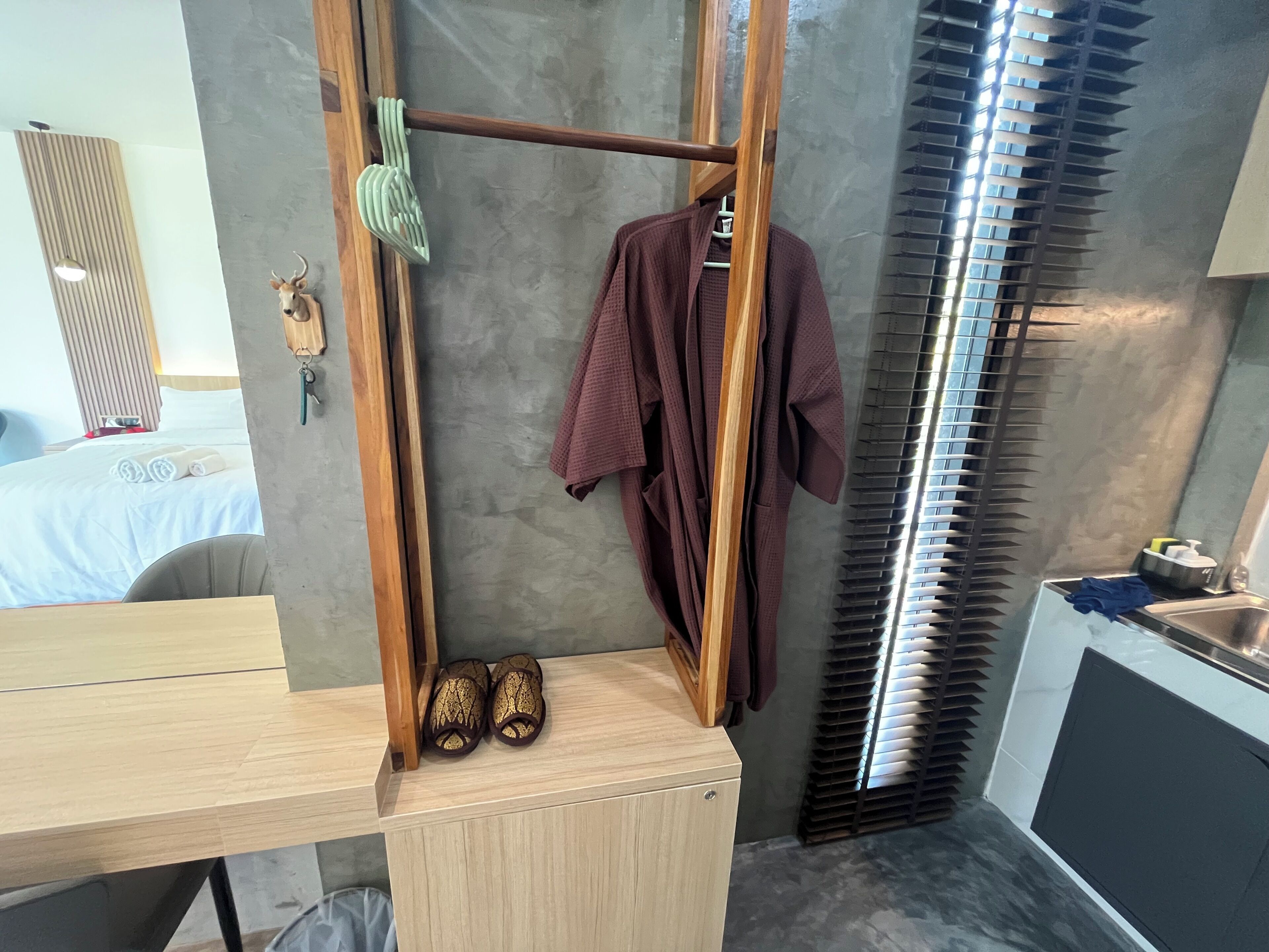 A hotel room with a wooden desk, a hanging robe, a pair of slippers, a wall-mounted towel rack, and a window with blinds.