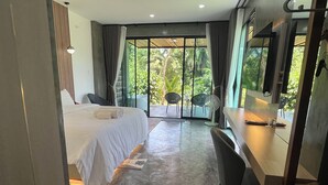 A modern hotel room with a bed, a desk, a chair, and a large window with a view of greenery. - Darisa Home (Surat Thani)
