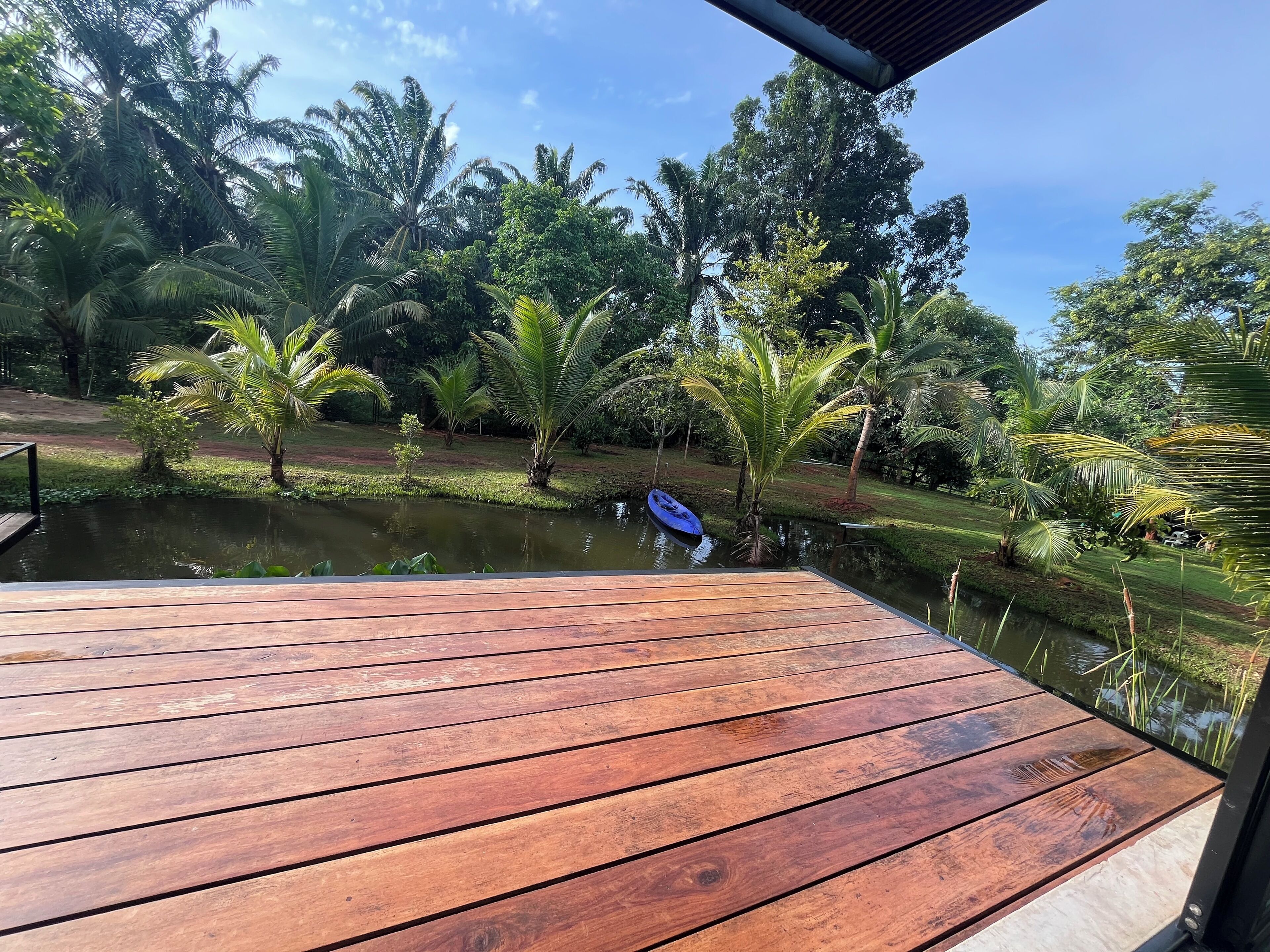 Eine Holzterrasse mit Blick auf einen Teich, auf dem ein blaues Kajak liegt, umgeben von üppigem Grün und Bäumen.