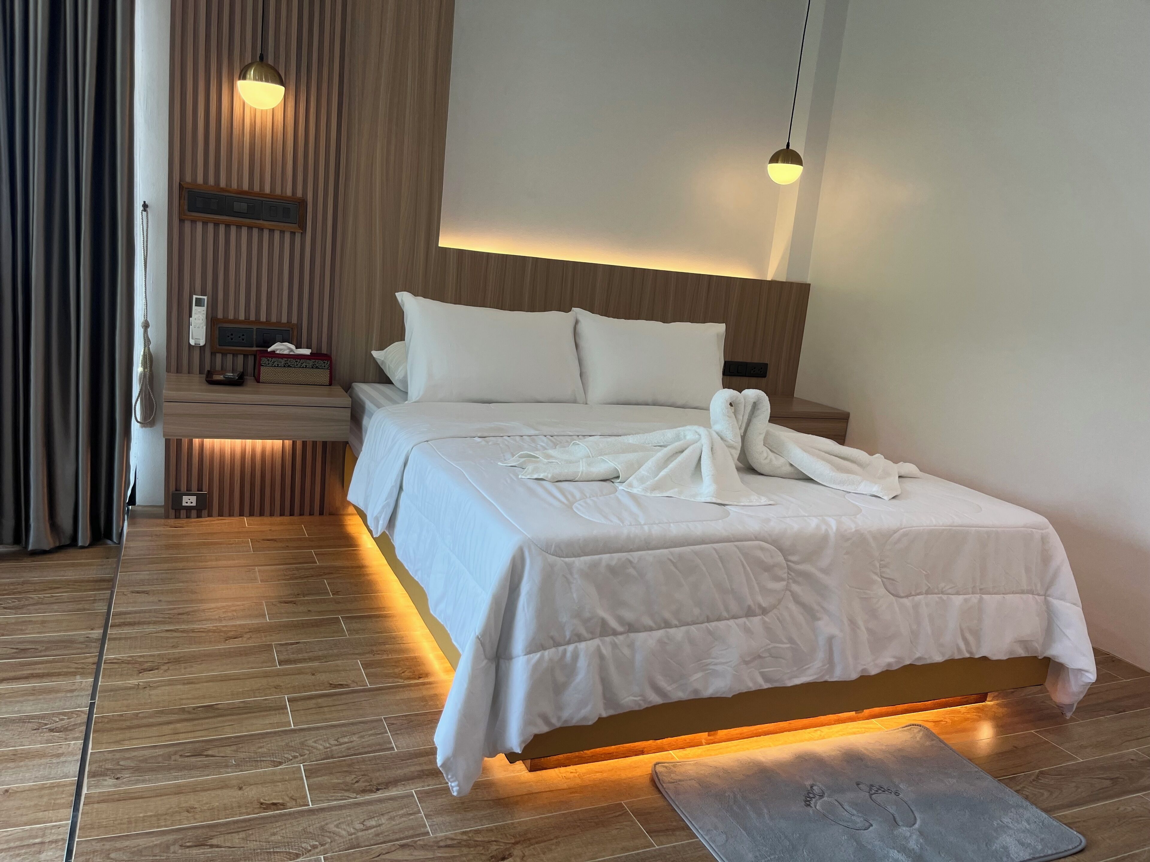 A hotel room with a wooden floor, a bed with white bedding, a bedside table with a lamp, and a wall-mounted panel.