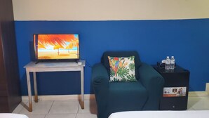 Ein Zimmer mit Fernseher auf einem Holztisch, einem grünen Sessel mit bunten Kissen und einer schwarzen Kommode mit Wasserflaschen.