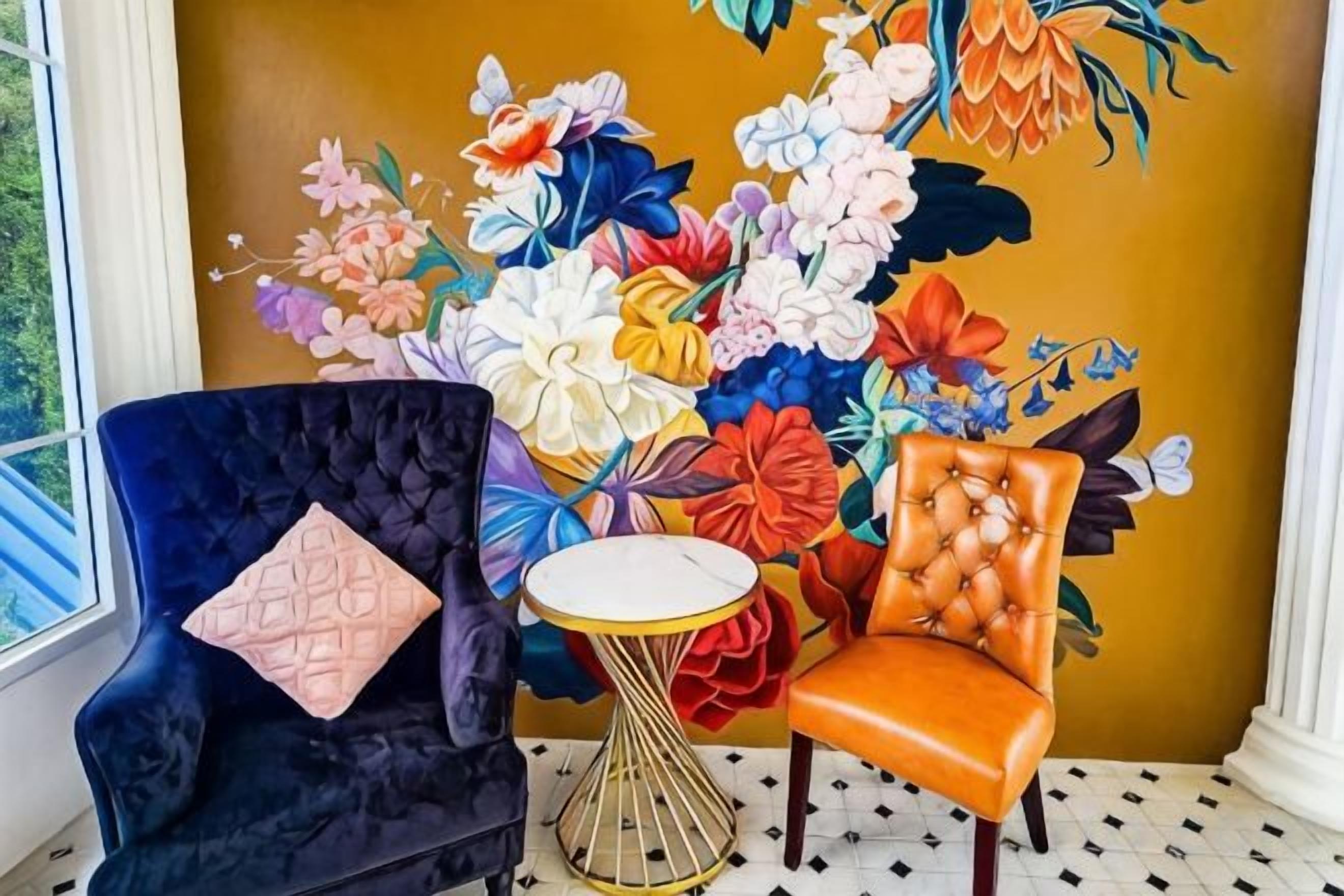 Una habitación con un amplio mural floral, un sillón azul oscuro con un cojín rosa y un sillón naranja con una mesita dorada.