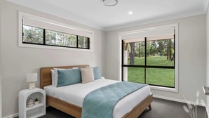 Une chambre à coucher comprenant un lit, une table de chevet, une lampe, une étagère avec des objets décoratifs et de grandes fenêtres donnant sur des arbres et de la verdure.