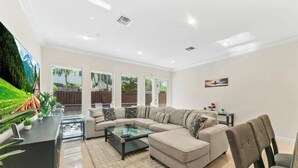 A spacious living room with a large sectional sofa, a glass coffee table, and a flat-screen TV mounted on the wall.