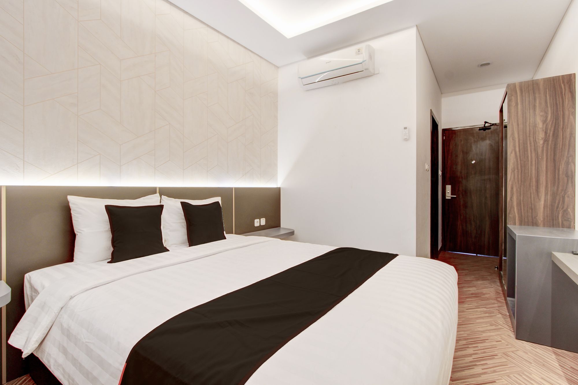 A modern hotel room with a large bed, a nightstand, an air conditioning unit, and a wooden door.