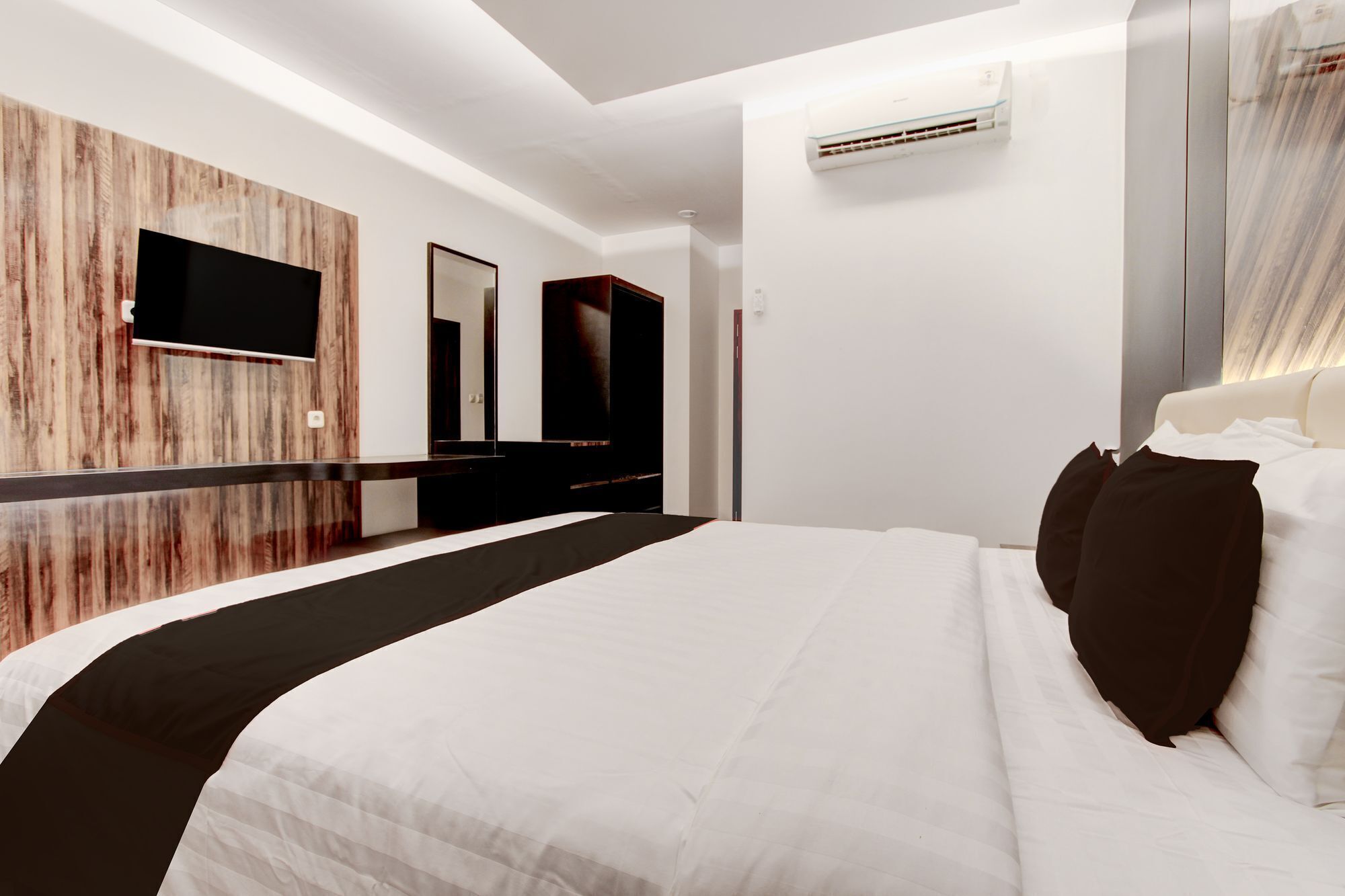 A modern hotel room with a large bed, a flat-screen TV mounted on a wooden wall, a mirror, and an air conditioning unit.
