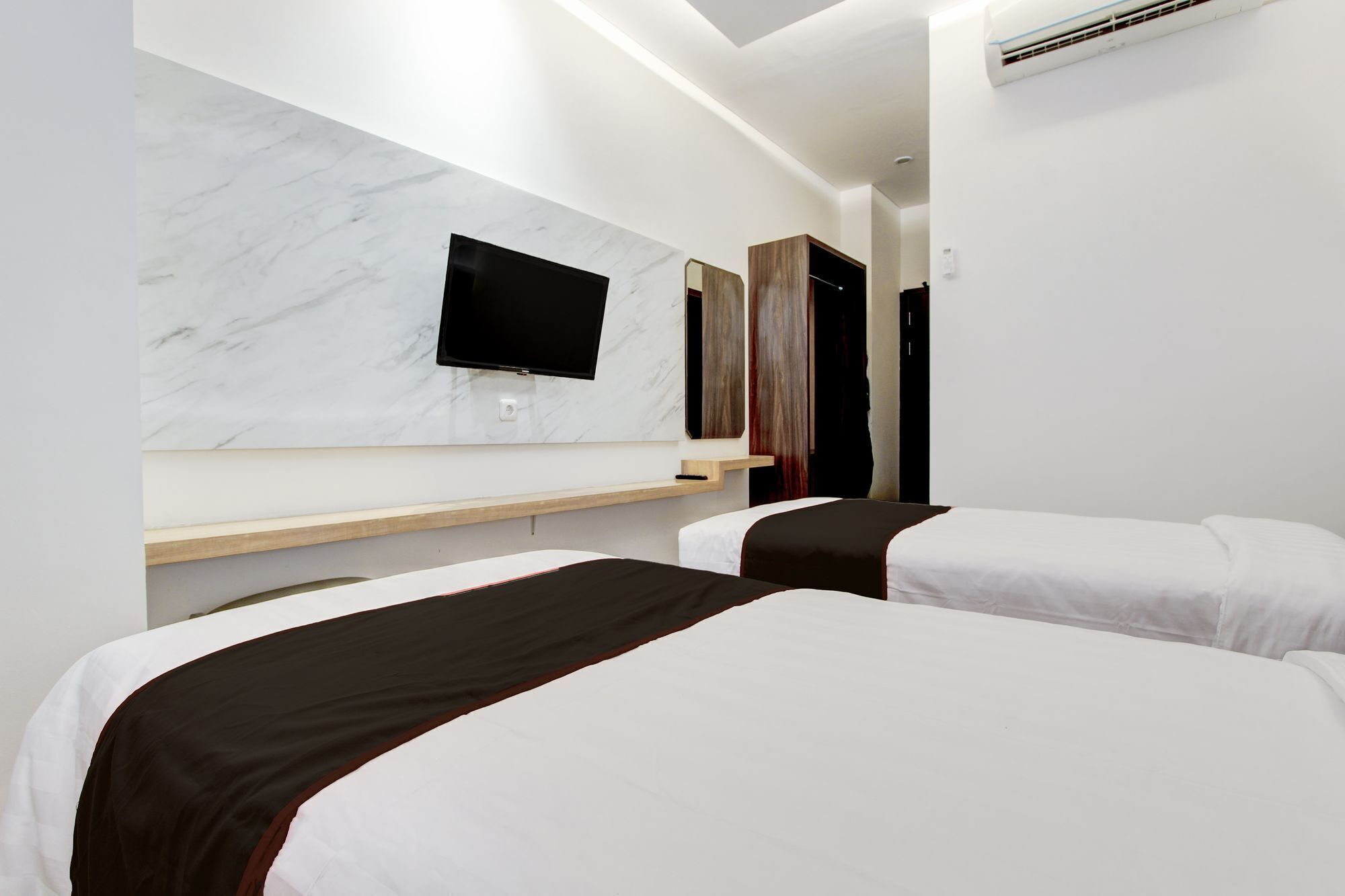 A hotel room with two beds, a flat-screen TV mounted on a marble wall, and an air conditioning unit.