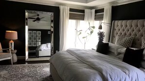 A modern bedroom with a large bed, a ceiling fan, a mirror, and a view of a cityscape through the window.