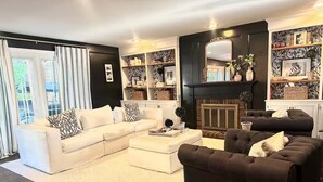 A living room with a white sectional sofa, a dark brown armchair, and a fireplace. There are shelves with decorative items and a large mirror above the fireplace.