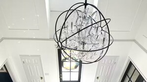 A spacious room with a large chandelier, wooden flooring, and a view through a doorway.