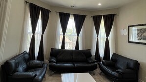 Ein Wohnzimmer mit drei schwarzen Ledersofas, einem weißen Couchtisch und großen Fenstern mit dunklen Vorhängen.