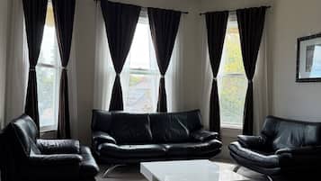 Ett modernt vardagsrum med en svart lädersoffgrupp, ett vitt soffbord och ett stort fönster med gardiner.