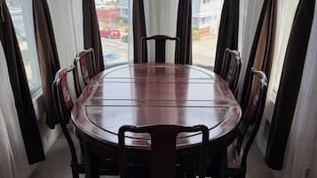 A dining area with a round table and chairs, large windows with curtains, and a view of a residential area.