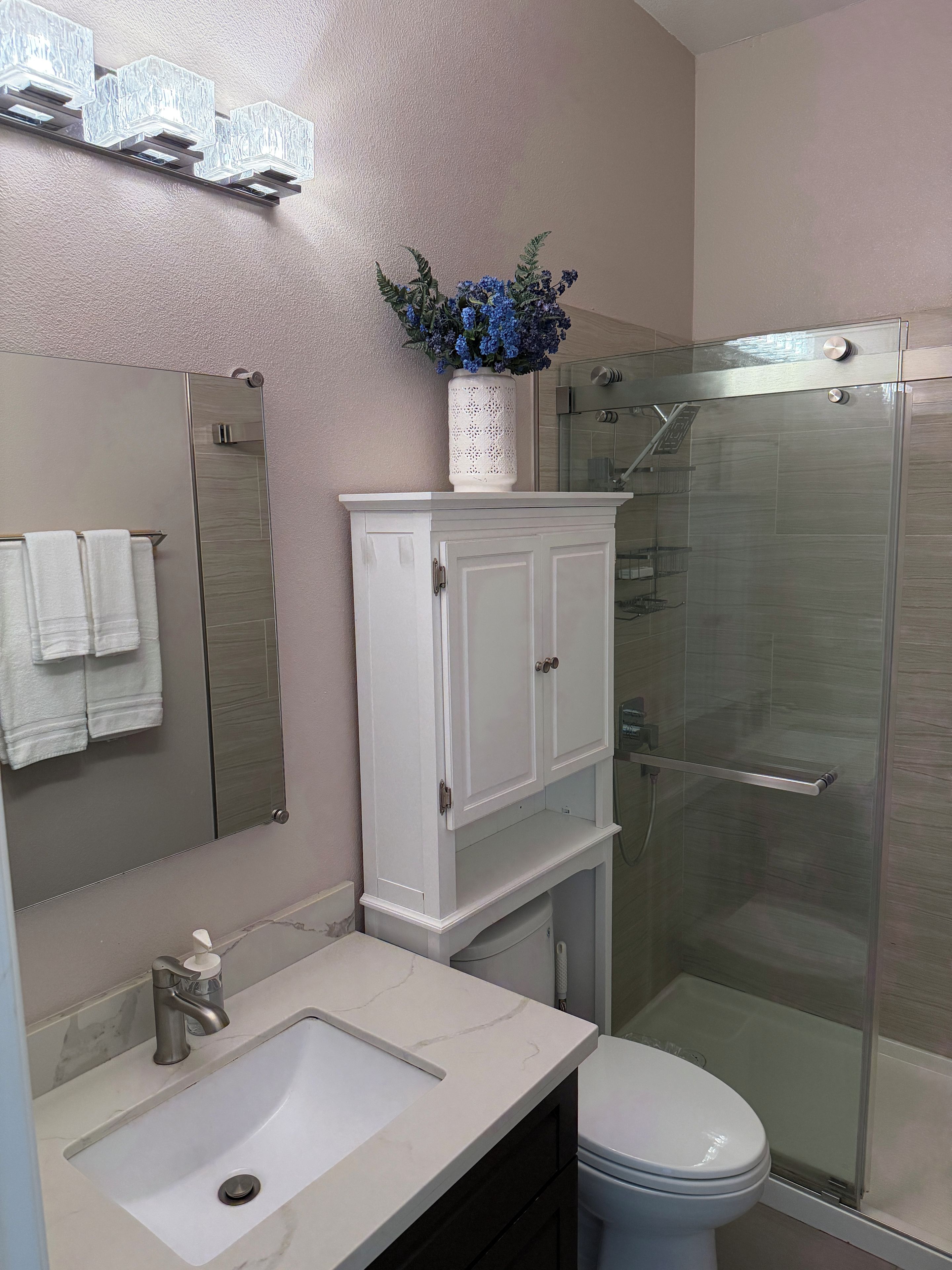 Un baño con tocador blanco, espejo, lavabo, inodoro y cabina de ducha con mampara de vidrio.