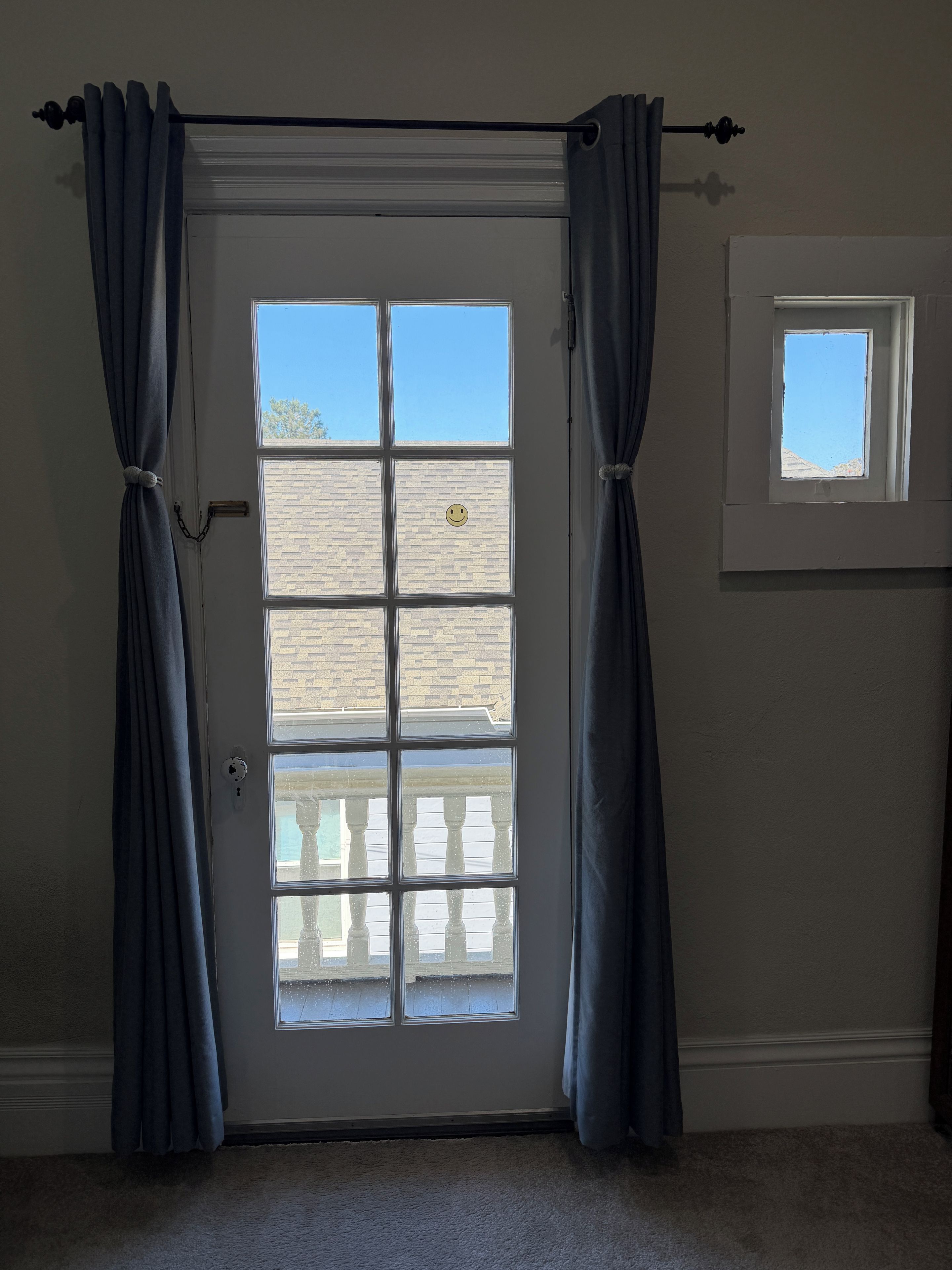 Una habitación con una puerta que tiene un ventanal con un adhesivo de carita sonriente, cortinas y un pequeño ventanuco encima.