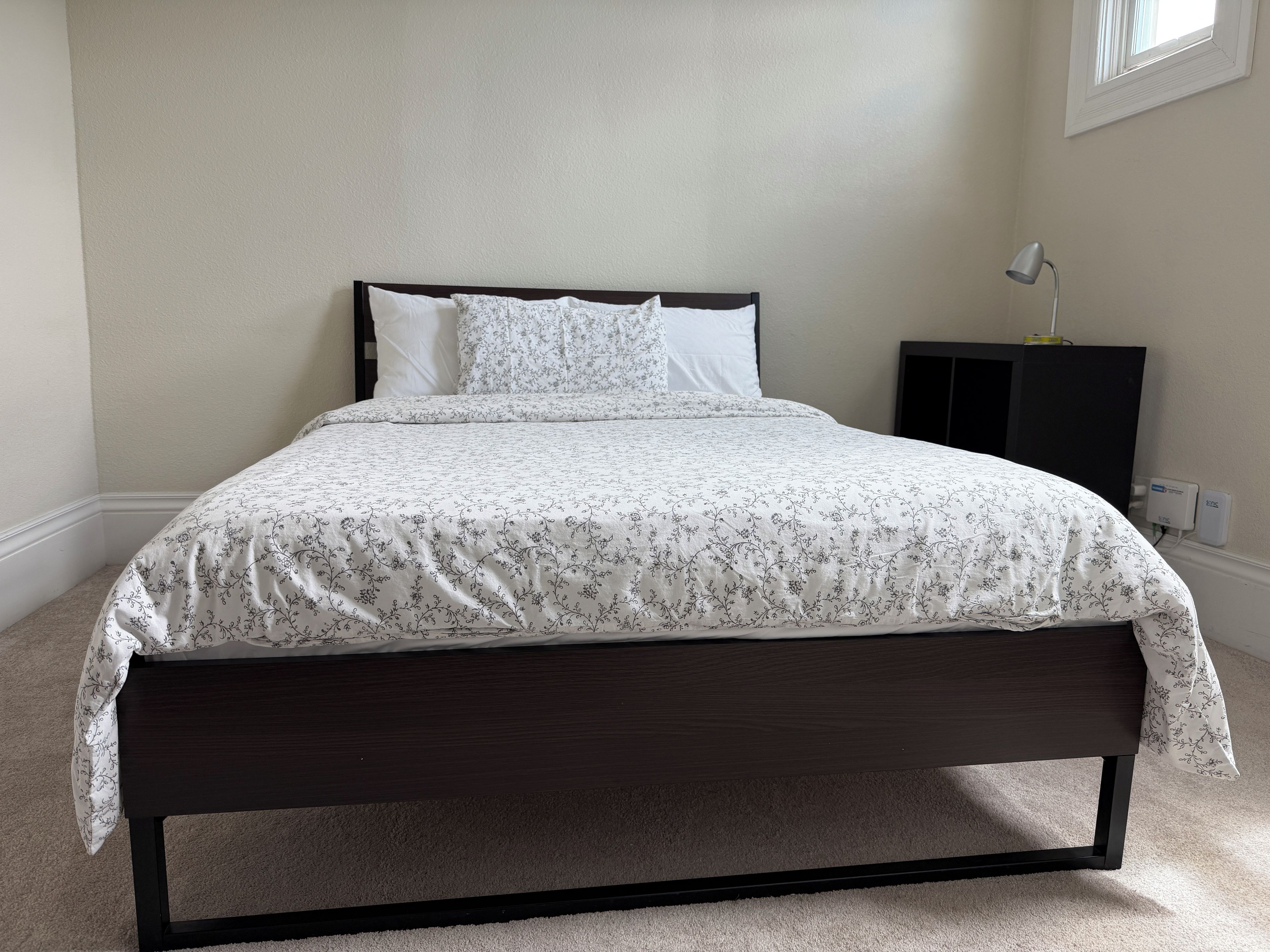 Una cama con estructura oscura, ropa de cama blanca con estampado floral, una mesita de noche con una lámpara y un enchufe en la pared.