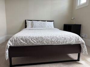 A bed with a dark frame, white bedding with a floral pattern, a bedside table with a lamp, and a power outlet on the wall. - Alameda Modern Victorian 4BD unit A (Alameda)