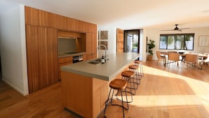 A modern kitchen with wooden cabinetry, a central island with bar stools, and a dining area with a large window. - Byron Oakland Oasis (Byron Bay)