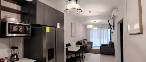 Eine moderne Küche mit integriertem Mikrowellenherd, Kühlschrank und Kaffeemaschine. Es gibt einen Essbereich mit Tisch und Stühlen, und eine Couch im Hintergrund.
