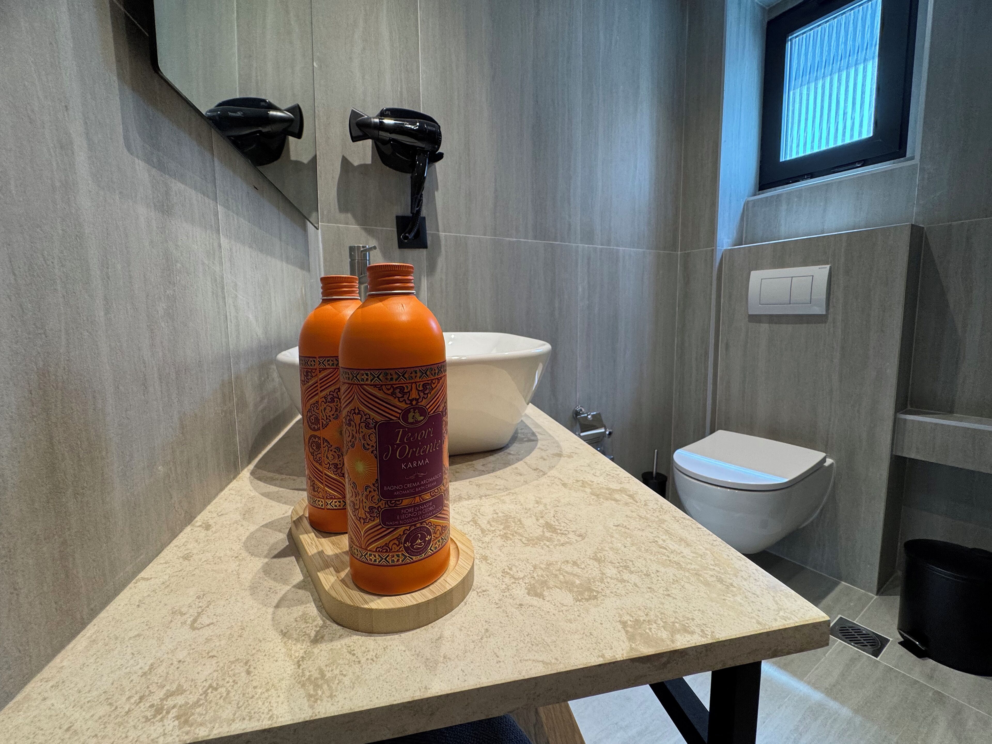 Une salle de bain avec un plan de toilette en marbre, deux flacons de la marque « Tesori d’Oriente Karma » qui y sont posés, un lavabo blanc et des toilettes.