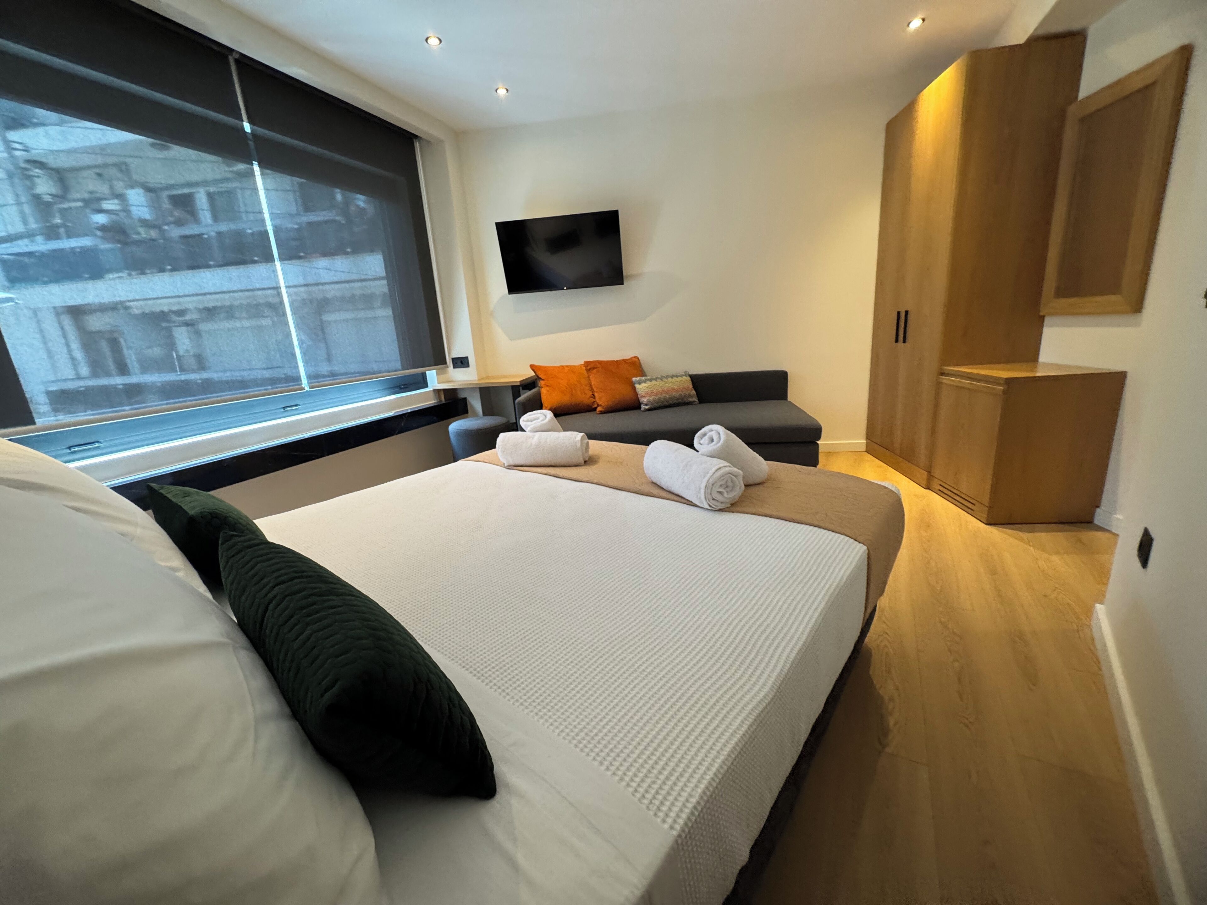 A modern hotel room with a large bed, a sofa, a TV mounted on the wall, and a wooden wardrobe.
