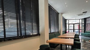 A modern indoor seating area with long tables and green cushioned benches, large windows with blinds, and a marble floor. - Downtown Suites (Thessaloniki)