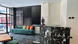 A modern reception area with a marble reception desk, a green sofa with cushions, and a view of the street through large windows. - Downtown Suites (Thessaloniki)