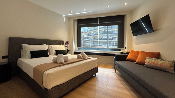 A hotel room with a large bed, a sofa, a TV, and a window with a city view. - Downtown Suites (Thessaloniki)