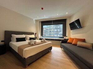 A hotel room with a large bed, a sofa, a TV, and a window with a city view. - Downtown Suites (Thessaloniki)