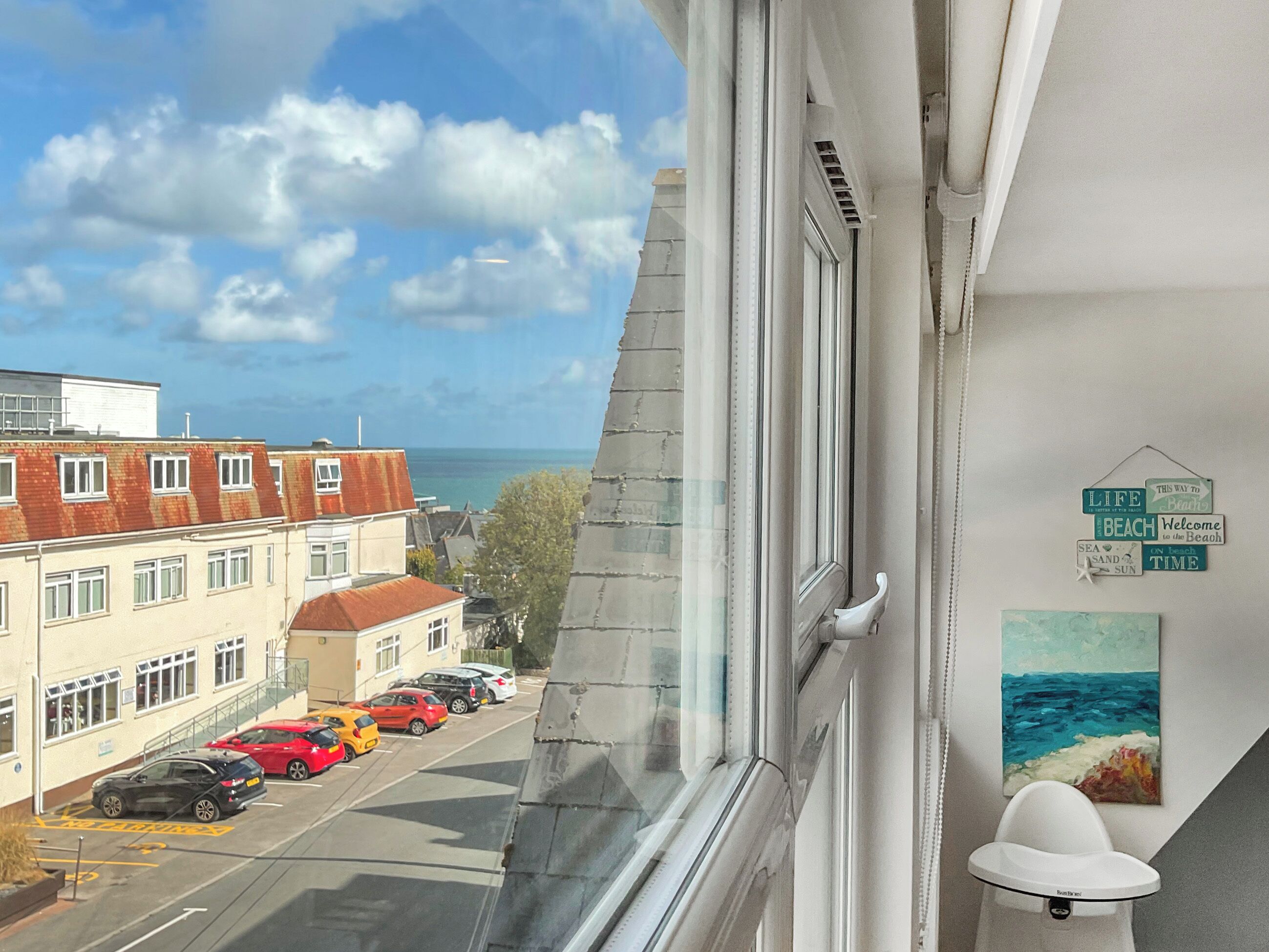 Blick aus einem Fenster auf eine Straße mit parkenden Autos, ein Gebäude mit rotem Dach und ein Schild mit Text zum Thema Strand.