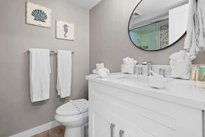 A bathroom with a toilet, white vanity, and a round mirror. There are two towels hanging on the wall and a seahorse picture. - Cozy Ocean Views at Beach and Tennis 4206 (Bonita Springs)