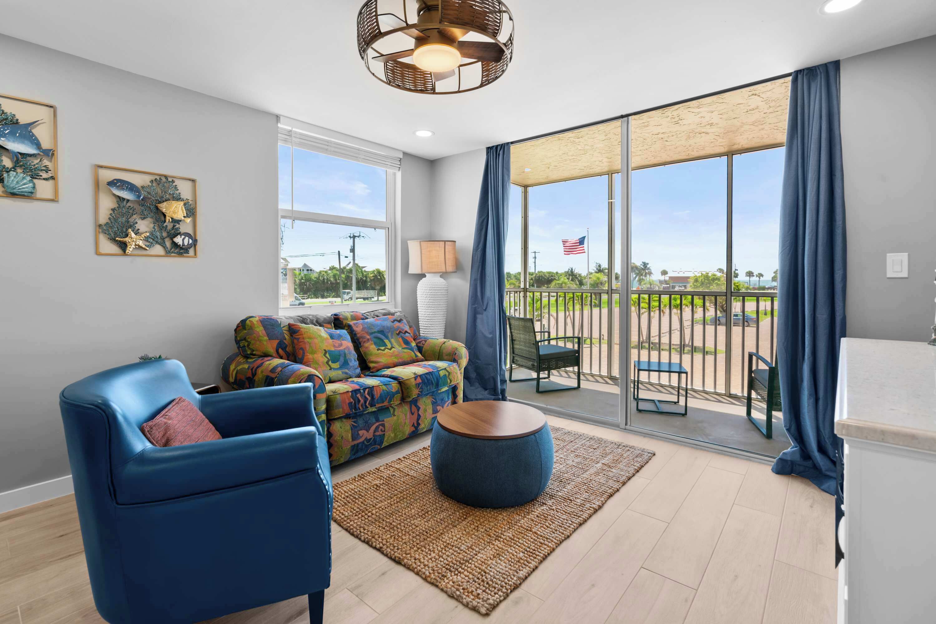 A modern living room with a blue sofa, a colorful patterned sofa, a round wooden coffee table, and a large sliding glass door leading to an outdoor patio.