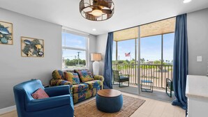 A modern living room with a blue sofa, a colorful patterned sofa, a round wooden coffee table, and a large sliding glass door leading to an outdoor patio.