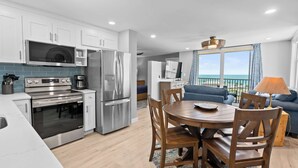 A modern kitchen with stainless steel appliances, a dining area with wooden chairs, and a view of the ocean through large windows. - Spectacular Ocean Views at Beach and Tennis 4705 (Bonita Springs)